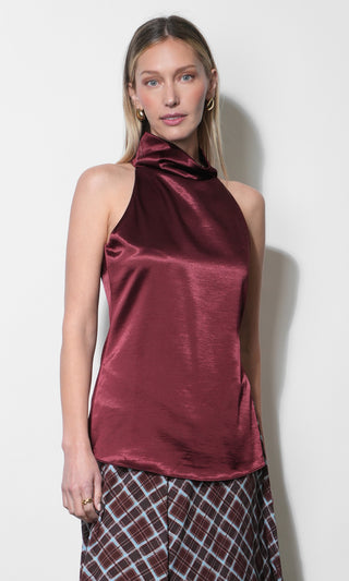 A woman modeling a sleeveless, high-neck satin top in a deep burgundy color, paired with a brown and white plaid skirt, against a plain off-white background.