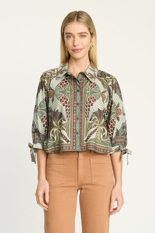 A woman modeling a cropped button-up blouse with a detailed botanical and paisley print in green, beige, and brown tones, paired with tan pants, against a plain light background.