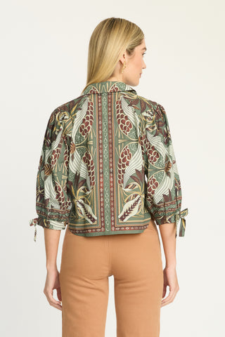 A woman shown from the back wearing a cropped button-up shirt with a detailed botanical and bird pattern in olive green, brown, and cream, paired with tan pants, against a plain white background.