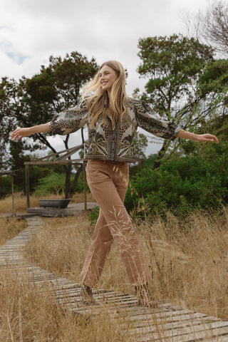 A woman stands on a wooden boardwalk in a grassy outdoor setting, wearing a patterned beige and black long-sleeve top with tan pants.