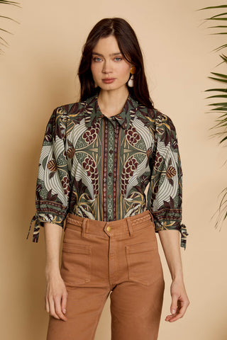 A woman modeling a long-sleeve button-up shirt with a detailed botanical and bird pattern in earthy tones, paired with brown pants, set against a warm beige background with soft plant shadows.