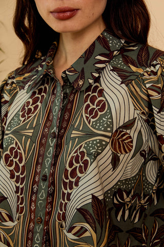 A close-up of a woman wearing a collared button-front blouse with an intricate botanical and abstract pattern in muted green, cream, brown, and burgundy tones.