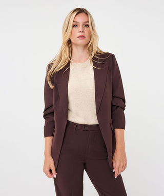 A blonde woman modeling a dark brown tailored blazer and matching trousers over a cream knit top, photographed against a clean light gray studio background.
