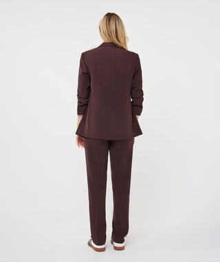 A woman viewed from the back wearing a matching dark plum-brown blazer and trousers set, standing against a plain light gray studio background.