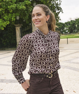A woman wearing a brown and beige geometric patterned long-sleeve blouse and dark brown pants, standing outdoors in a park-like setting with trees and a paved walkway.
