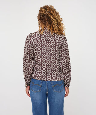 A woman viewed from the back wearing a brown and beige geometric patterned blouse with long sleeves and blue jeans, against a plain light gray studio background.