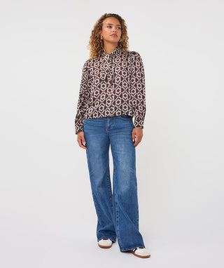 A woman modeling a long-sleeve button-up blouse with a brown, beige, and white geometric pattern, paired with wide-leg blue jeans and white sneakers, against a plain light gray studio background.