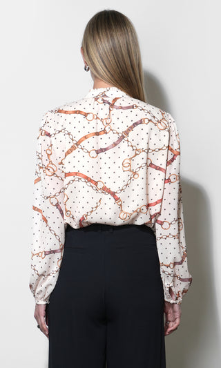 A woman viewed from the back wearing a long-sleeve blouse with an allover horsebit and chain print over a white base with small black polka dots, paired with black trousers.