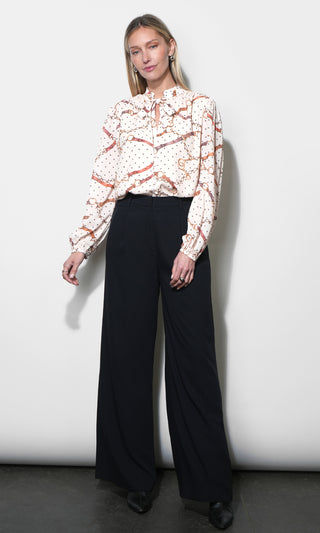 A woman modeling a long-sleeve blouse with a cream base, orange-brown abstract pattern, and small white dots, paired with black wide-leg trousers, standing against a light gray studio wall.