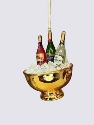 A gold champagne bucket ornament filled with white faux ice and three miniature champagne bottles, hanging against a neutral gray background.