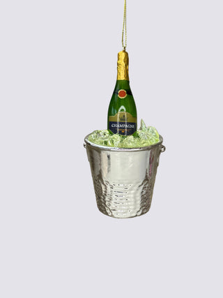 A miniature champagne bottle nestled in clear ice inside a hammered silver bucket, suspended against a neutral beige background.