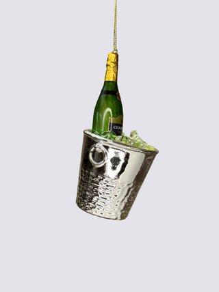 A hammered silver metal champagne bucket ornament filled with green ice and a miniature green champagne bottle, suspended by a gold string against a neutral beige background.