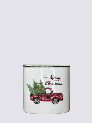 A white enamel-style vase with a gold rim, featuring a vintage red pickup truck carrying a green Christmas tree and the words 'Merry Christmas' printed on the front, set against a neutral beige fabric backdrop.