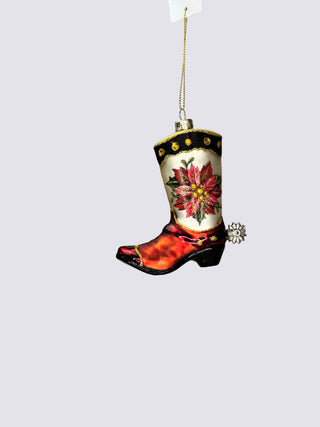 A hand-painted Christmas ornament shaped like a cowboy boot, featuring a red and orange flame pattern on the lower half, a white upper with a red poinsettia flower, and black trim with gold dots. It hangs from a gold string against a neutral gray background.