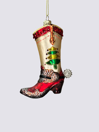 A Christmas ornament shaped like a cowboy boot, featuring a cream shaft with a green and gold Christmas tree motif, red sequined trim, a red and silver glittery pointed toe, and a small silver flower ornament hanging from a gold string against a neutral gray background.