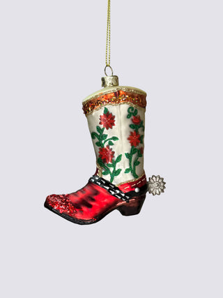 A hand-painted glass Christmas ornament shaped like a cowboy boot, featuring a red glittery hoof, a cream shaft with red flowers and green leaves, and a gold hanging string against a neutral gray background.
