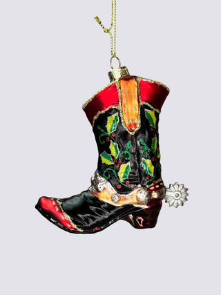 A hand holds a hand-painted Christmas ornament shaped like a western cowboy boot. The boot is primarily black with red trim, green and gold embroidered details, and a silver-toned flower ornament hanging from the heel.