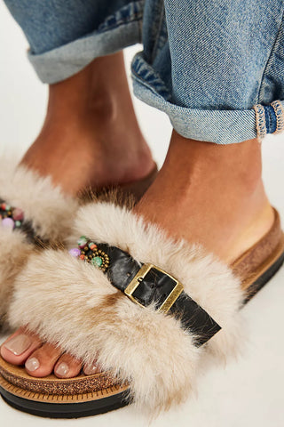  Fluffy beige faux-fur slide sandals with black straps, a gold-tone buckle, and colorful bead accents, paired with cuffed blue jeans.