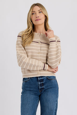 A woman modeling a beige and white striped knit cardigan with dark brown trim, paired with blue jeans, against a light gray studio background.