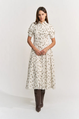A woman modeling a short-sleeved midi dress with a brown and beige botanical print on a cream background, paired with dark brown knee-high boots, against a plain white studio background.