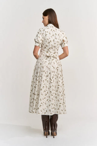 A woman viewed from the back wearing a cream-colored midi dress with a brown floral print, short puff sleeves, and a tie waist, paired with dark brown boots against a plain white studio background.