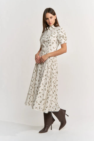 A woman modeling a short-sleeved midi dress with a white base and a delicate brown floral print, paired with dark brown heeled ankle boots, against a plain light gray studio background.