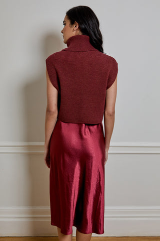 A woman shown from the back wearing a sleeveless ribbed turtleneck sweater and a satin-like midi skirt in a deep wine red color, set against a neutral indoor wall.