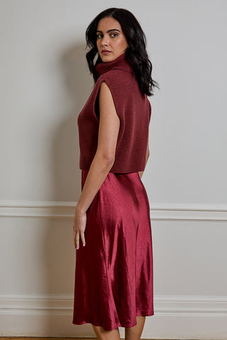A woman wearing a sleeveless ribbed knit turtleneck top and a matching satin midi skirt in a deep burgundy red, standing against a neutral off-white wall.