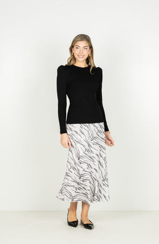 A woman modeling a black long-sleeve knit top paired with a white midi skirt featuring a gray abstract brushstroke pattern, standing against a plain white studio background.