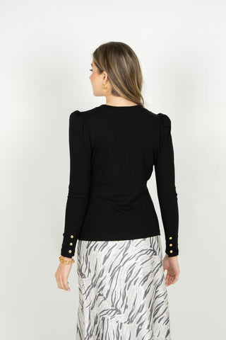 A woman shown from the back wearing a fitted black long-sleeve top with gold button details on the cuffs and a white skirt with a gray abstract pattern, against a plain light background.