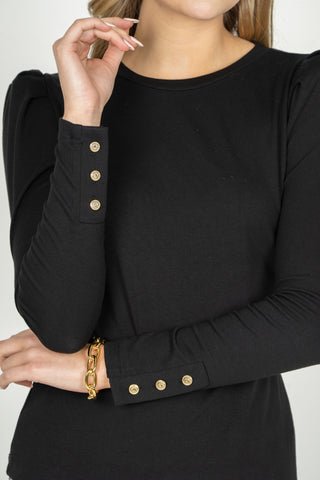 A close-up of a woman wearing a black long-sleeve top with gold button details on the cuffs, set against a plain light gray background.