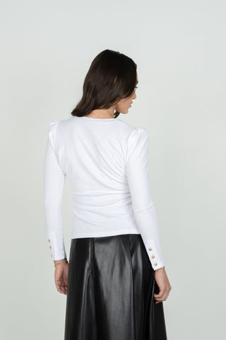 A woman shown from the back wearing a fitted white long-sleeve top with gold button details on the cuffs and a black skirt, against a plain light gray studio background.