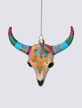 A hand-painted bull skull Christmas ornament with colorful floral and butterfly designs, hanging from a gold string against a neutral background.