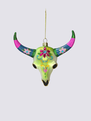 A hand holds a decorative bull skull ornament with bright green, pink, blue, and gold painted details against a neutral beige background.