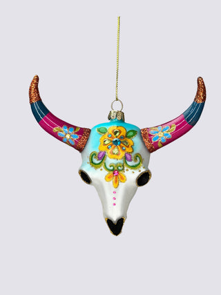 A hand holds a hand-painted bull skull Christmas ornament with colorful floral designs and curved horns against a neutral gray background.