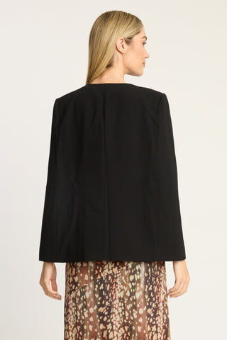 A woman shown from the back wearing a black long-sleeve cropped jacket with a gathered center back seam, paired with a brown and beige patterned skirt, against a plain light background.