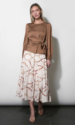 A woman modeling a long-sleeve brown satin top with a twisted waist detail and a white midi skirt featuring a rust-orange abstract pattern and small gold dots, set against a plain light gray wall.