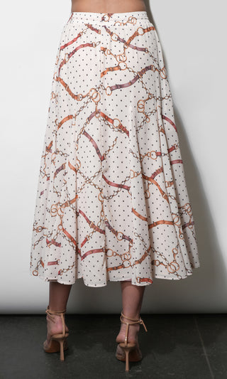 A woman's A-line midi skirt with an allover equestrian-inspired pattern of gold chain links, brown leather straps, and small black polka dots on an off-white background. The skirt is shown from the waist down on a model wearing brown strappy heels against a plain gray wall.