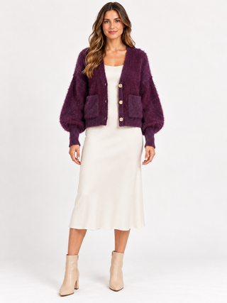 A woman modeling a fuzzy dark purple cropped cardigan with gold buttons and front pockets, worn over a white midi dress and paired with beige ankle boots, against a plain white studio background.