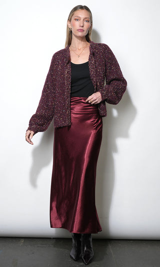 A woman models a burgundy tweed jacket with voluminous sleeves, worn over a black top and a matching satin midi skirt, paired with black pointed-toe boots against a white studio background.