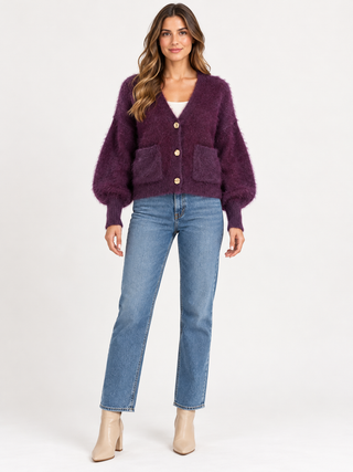 A woman modeling a fuzzy purple cardigan with front pockets and gold buttons, paired with blue jeans and beige ankle boots, against a plain white studio background.