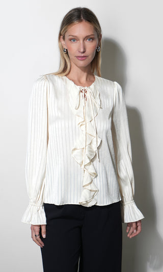 A woman modeling a cream-colored long-sleeve blouse with vertical pleats, a ruffled front tie detail, and ruffled cuffs, paired with black pants against a plain light gray background.