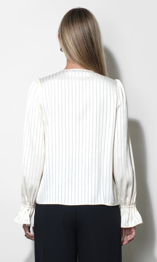 A woman viewed from the back wearing a white long-sleeve blouse with thin vertical pinstripes and ruffled cuffs, paired with black pants, against a plain light gray wall.