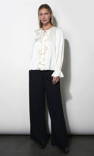 A woman modeling a white long-sleeve blouse with vertical pleats and a ruffled tie neckline, paired with black wide-leg trousers, standing against a plain light gray wall.