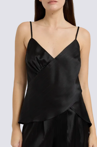 A close-up of a model wearing a black satin wrap-style camisole top with thin spaghetti straps and a draped, asymmetrical front overlay, paired with black trousers against a light gray studio background.