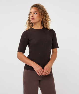 A woman with curly blonde hair wearing a dark brown short-sleeve knit top with subtle speckled details and matching brown pants, standing against a light gray studio background.