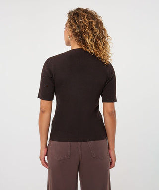 A woman with curly blonde hair is shown from the back wearing a dark brown short-sleeve knit top and brown pants against a light gray studio background.