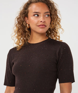 A woman with curly light brown hair wearing a dark brown ribbed short-sleeve top with subtle gold speckles, photographed against a light gray studio background.