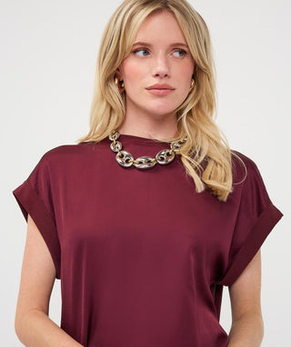 A blonde woman wearing a short-sleeve, boat-neck top in deep burgundy with a chunky gold chain necklace, photographed against a light gray studio background.