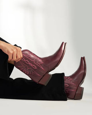 A person wearing burgundy leather cowboy boots with pink western-style stitching, shown from the hand down against a clean white studio background.